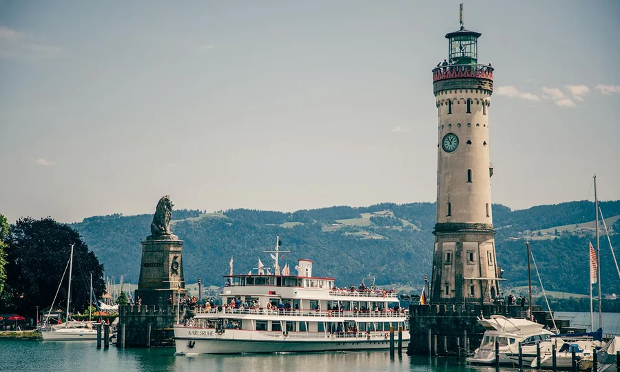 Blick auf den historischen Leuchtturm und die Hafeneinfahrt von Lindau mit einem Ausflugsschiff vor alpiner Kulisse