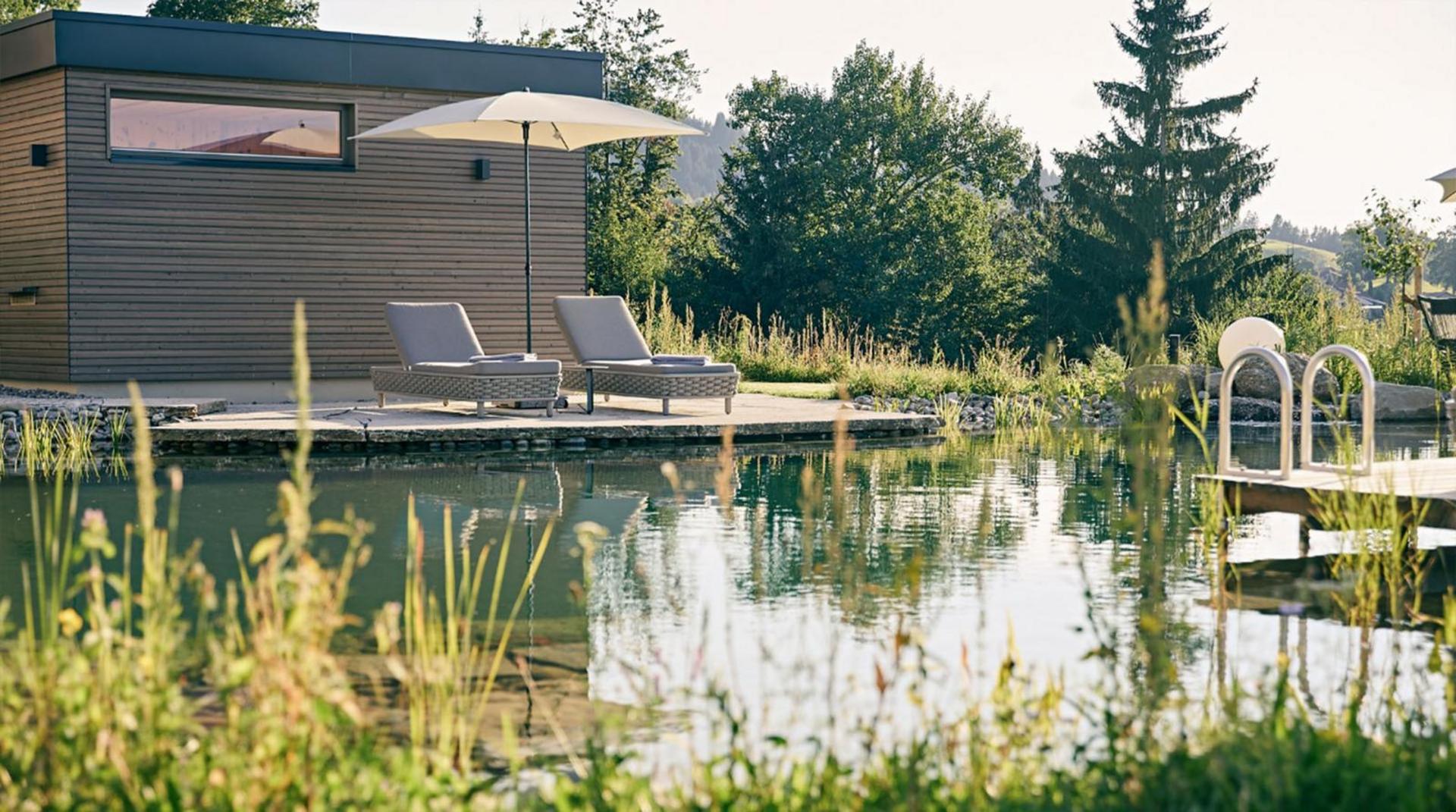 Zwei Liegen mit Sonnenschirm an einem Holzdeck direkt am Naturschwimmteich, umgeben von Grünpflanzen und Bäumen.