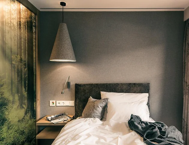 Bett mit grauem Kopfteil und Nachttisch, darüber große Hängelampe, ruhige Einrichtung in Naturtönen.