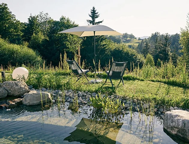 Zwei Gartenstühle mit Sonnenschirm am Rand des Naturschwimmteichs im Hotel Birkenhof, umgeben von Wiese, Steinen und Blick ins Grüne.