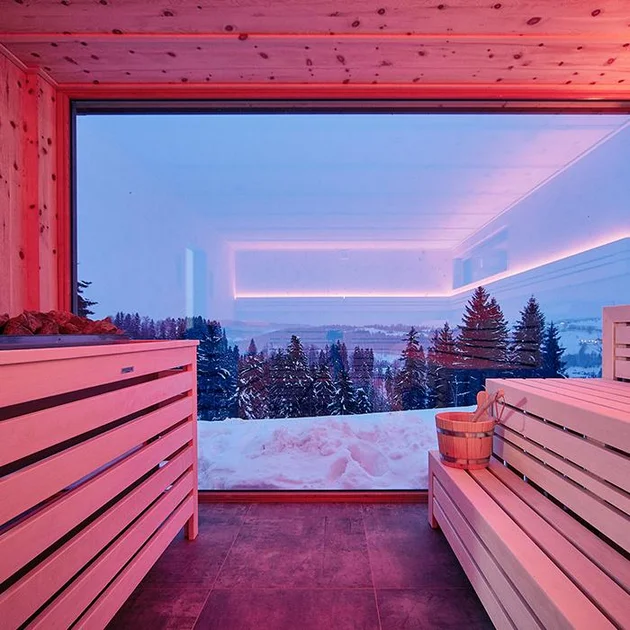 Moderne Sauna mit großem Panoramafenster und Blick auf die verschneite Winterlandschaft, stimmungsvoll beleuchtet.