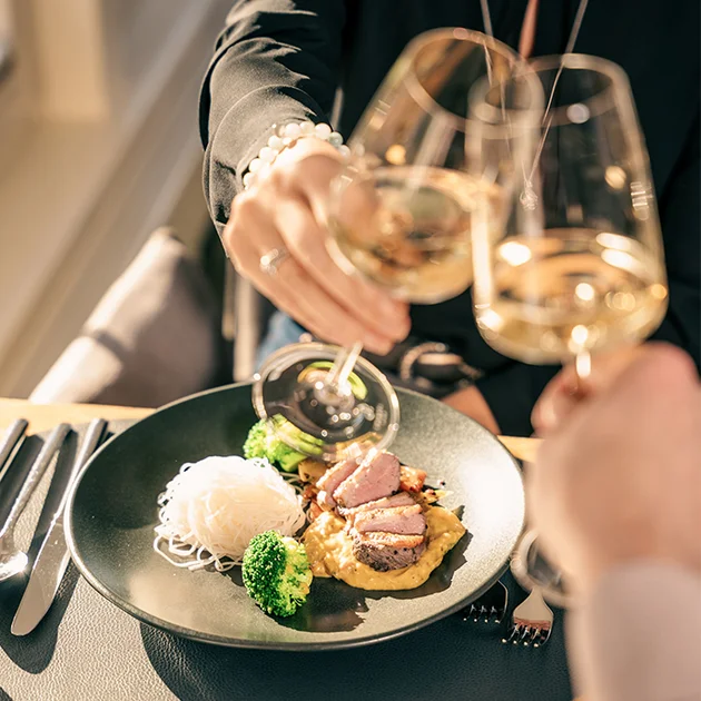 Zwei Gäste stoßen mit Weingläsern an auf dem Tisch steht ein Fleisch Gericht mit Brokkoli und Gemüse