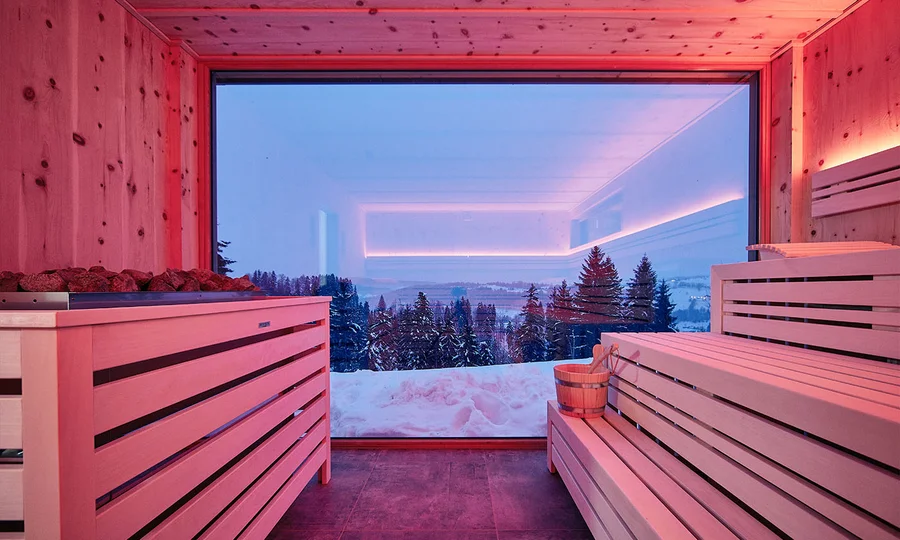 Holzsauna mit roten Lichteffekten und Blick auf verschneite Bäume und Berge.