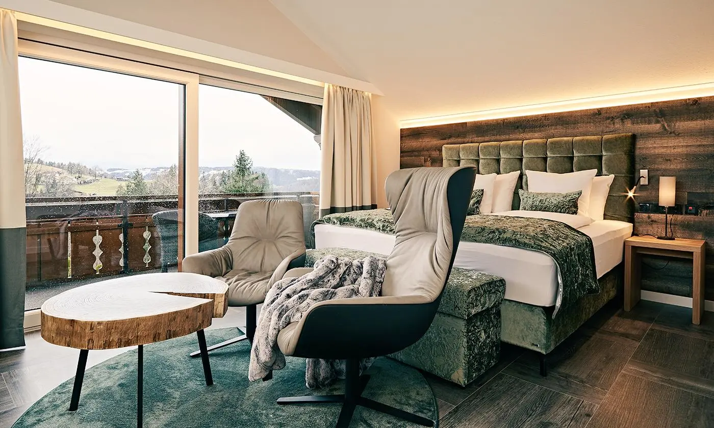 Stylish bedroom with green box-spring bed, armchairs and balcony view at Hotel Birkenhof Oberstaufen.