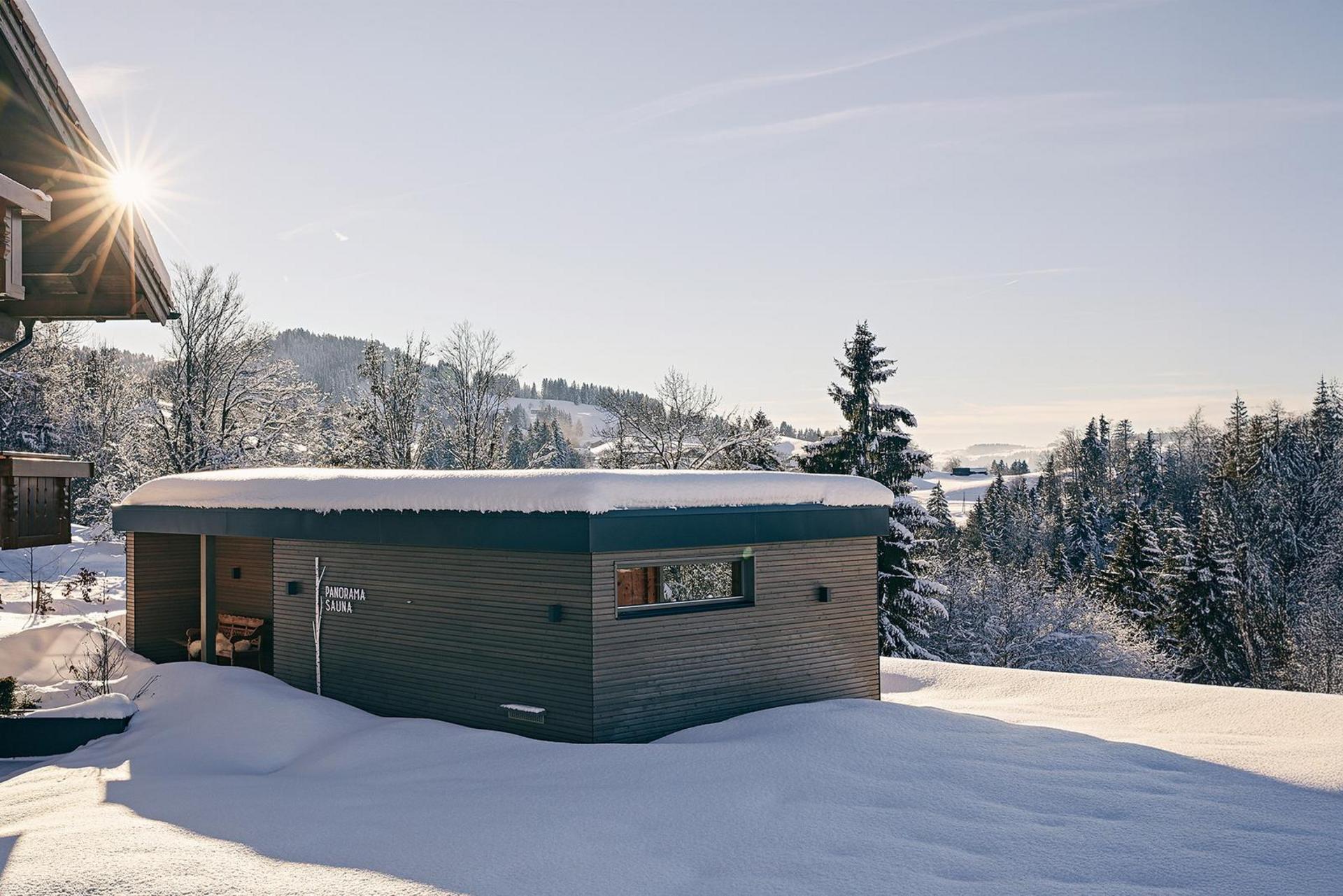 Panoramasauna2_Winter_Hotel_Birkenhof_Oberstaufen_Allgaeu Winteransicht des modernen Spa-Gebäudes mit schneebedecktem Dach und umliegender Winterlandschaft.