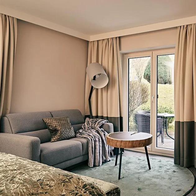 Helles Hotelzimmer mit Sofa, Stehlampe und Zugang zur Terrasse im Hotel Birkenhof Oberstaufen.