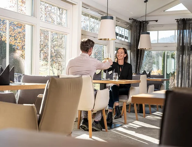 Paar stößt im hellen Wintergarten-Restaurant des Hotel Birkenhof bei Sonnenlicht an.