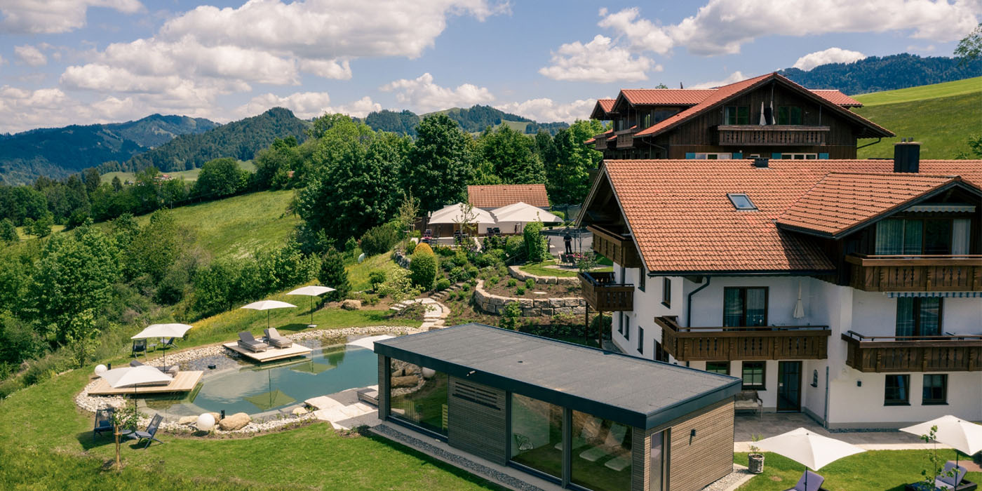 Außenansicht des Hotel Birkenhof Oberstaufen mit Natur-Schwimmteich, Liegebereich und Blick auf die umliegende Allgäuer Hügellandschaft.