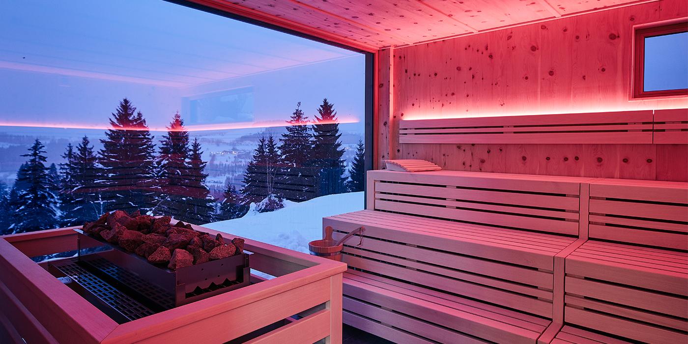 Moderne Sauna im Birkenhof mit roten Lichteffekten und weitem Blick auf verschneite Berglandschaft.