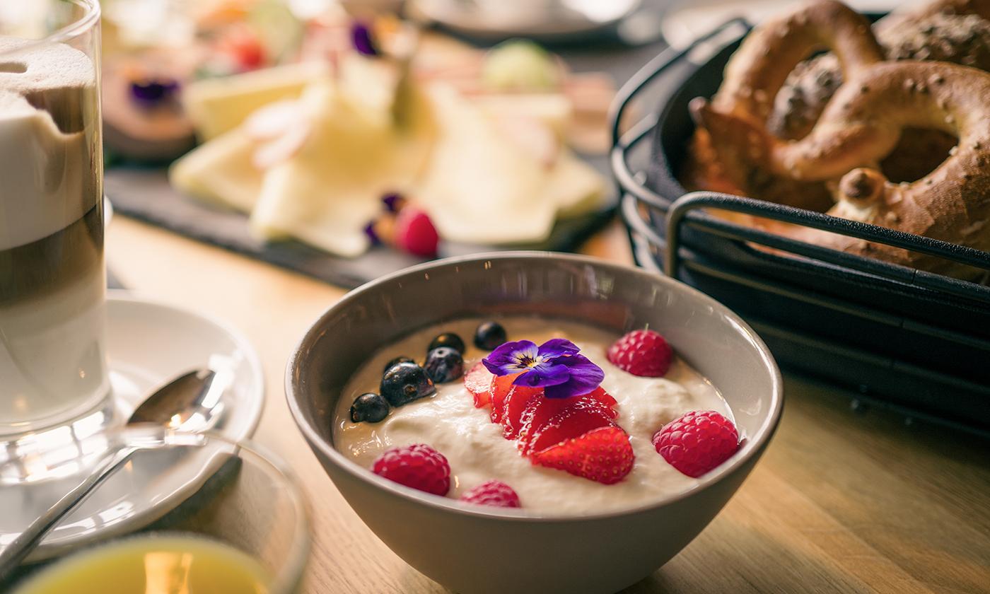 Schüssel mit Joghurt, Beeren und essbaren Blüten, daneben Brezeln, Kaffee und Obst.