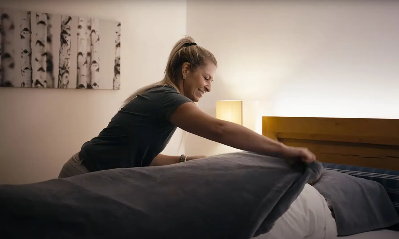 Frau legt einem Gast im Bett eine warme Decke über, gemütliches Zimmer mit Holzdetails und sanfter Beleuchtung.