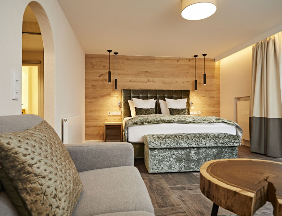 Gemütliches Doppelzimmer mit Boxspringbett, grünem Stoffbezug und Holzrückwand, Sofa im Vordergrund und rustikalem Couchtisch.